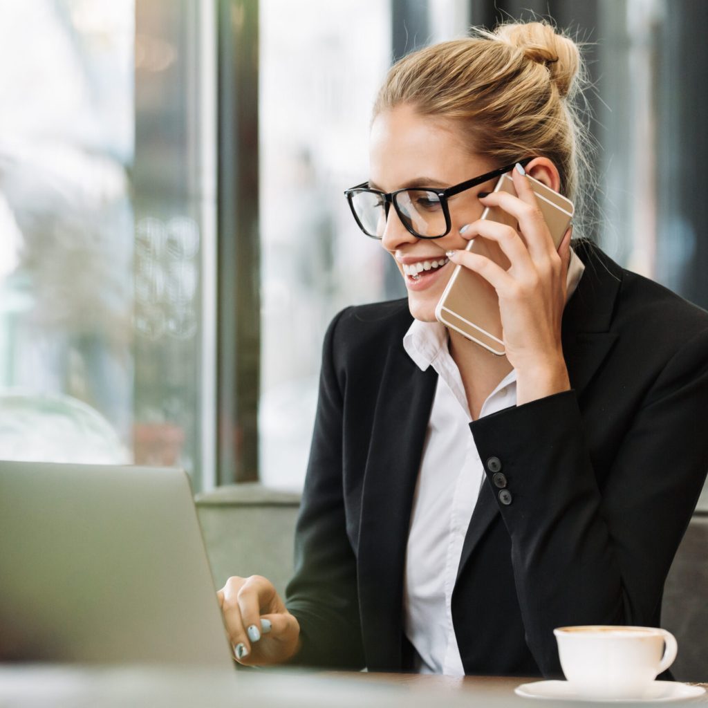 Photo of young happy smiling blonde business woman sitting indoors in cafe using laptop computer talking by mobile phone. Looking aside.